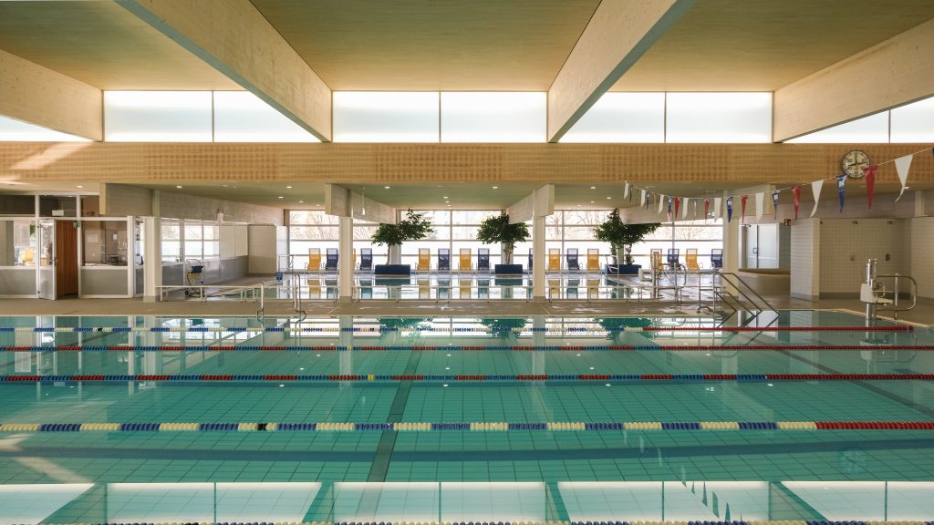 Sanierung des alten Hallenbades inklusive Ausbau mittels eines Hochdaches. Die Akustikverkleidungen in Fichte ergeben ein sehr ruhiges Gesamtbild. Holzverkleidungen sind wesentlich unempfindlicher gegen Chlordämpfe als Metallverkleidungen. Die Lichtnischen wurden mittels transluzenter Gläser luftdicht gegenüber den Chlordämpfen geschützt.